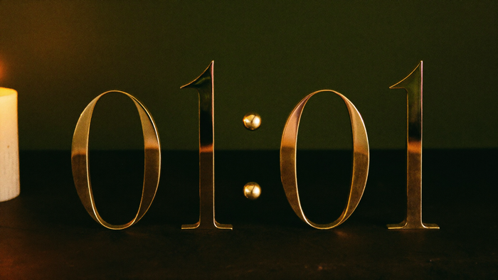 Brass numerals reading 01:01 on a dark stone ledge in front of a deep green wall, a lit candle at the left