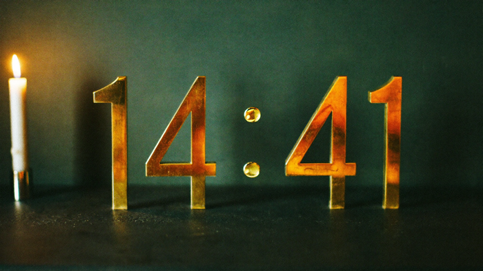 Large brass numerals 14:41 on dark stone in front of a deep green wall, with a lit white candle on the left