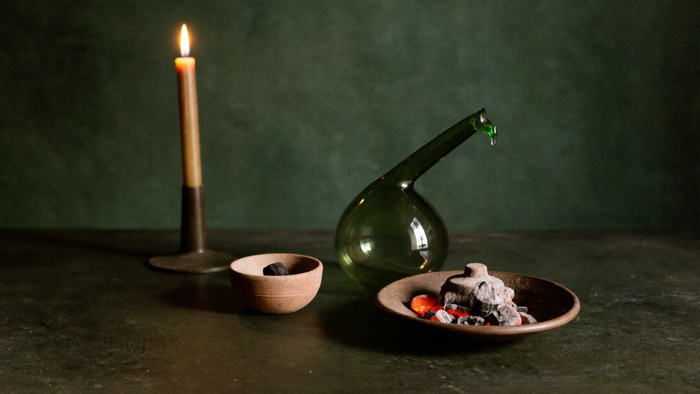 Green glass alchemical retort beside a clay dish of glowing coals and a lit taper candle on a dark stone table