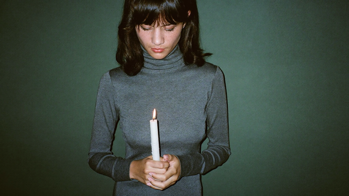 A woman in a plain grey turtleneck holding a lit white candle against a dark green wall