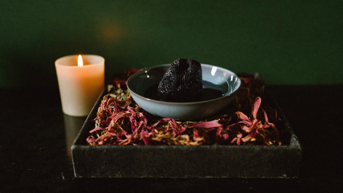 A candle, a bowl of dark water with a black stone and dried petals on a stone altar tray