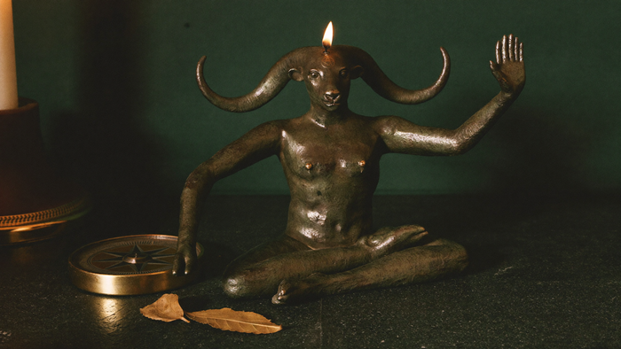 Bronze goat-headed figure seated cross-legged, one hand raised, a lit candle burning above its horns
