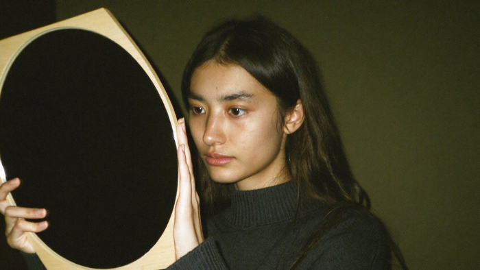 A young woman in a dark sweater holding up a round matte black scrying mirror in a wooden frame