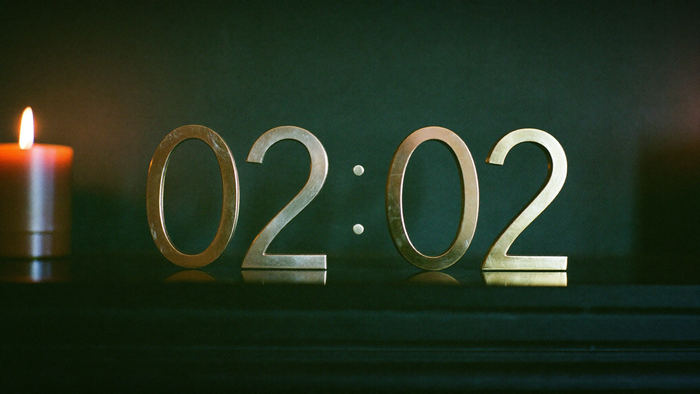 Brass numerals reading 02:02 on a dark shelf next to a lit orange candle against a deep green wall