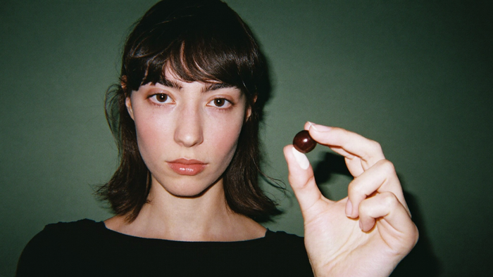 A young woman holding a single glossy brown buckeye seed between two fingers against a dark green wall