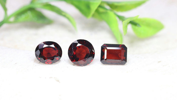 Three faceted deep red garnets, oval, round and emerald cut, on a pale stone surface with soft green leaves behind