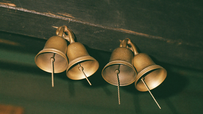 Four small brass bells hanging in two matched pairs from a dark wooden beam in warm low light