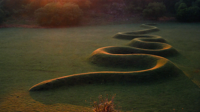 Grass-covered serpent-shaped earthwork winding across a meadow at dusk