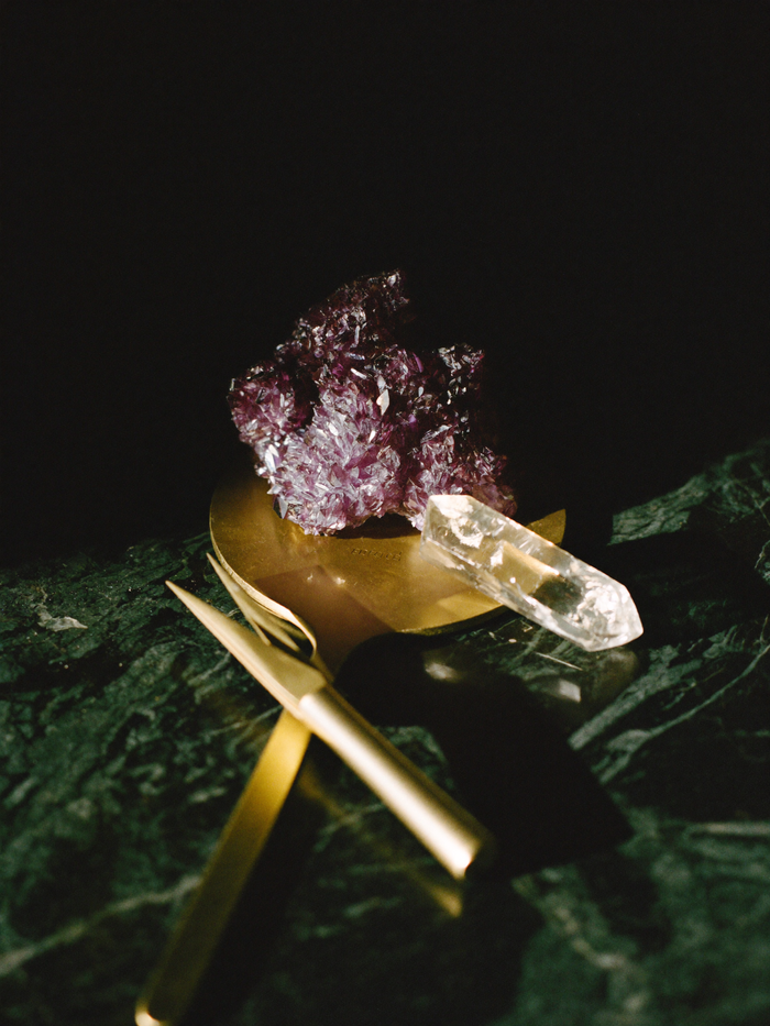 Raw amethyst cluster and clear quartz point beside a jeweller's loupe and brass tweezers on dark marble