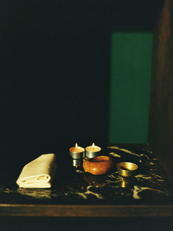 A small home altar with a candle and a few sacred objects on polished dark marble, lit by warm golden light against a deep emerald-green background.