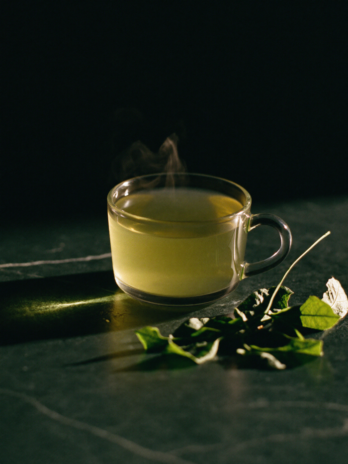 A glass cup of pale green tea steaming on polished dark marble, warm golden light glowing through it against a deep emerald-green background.