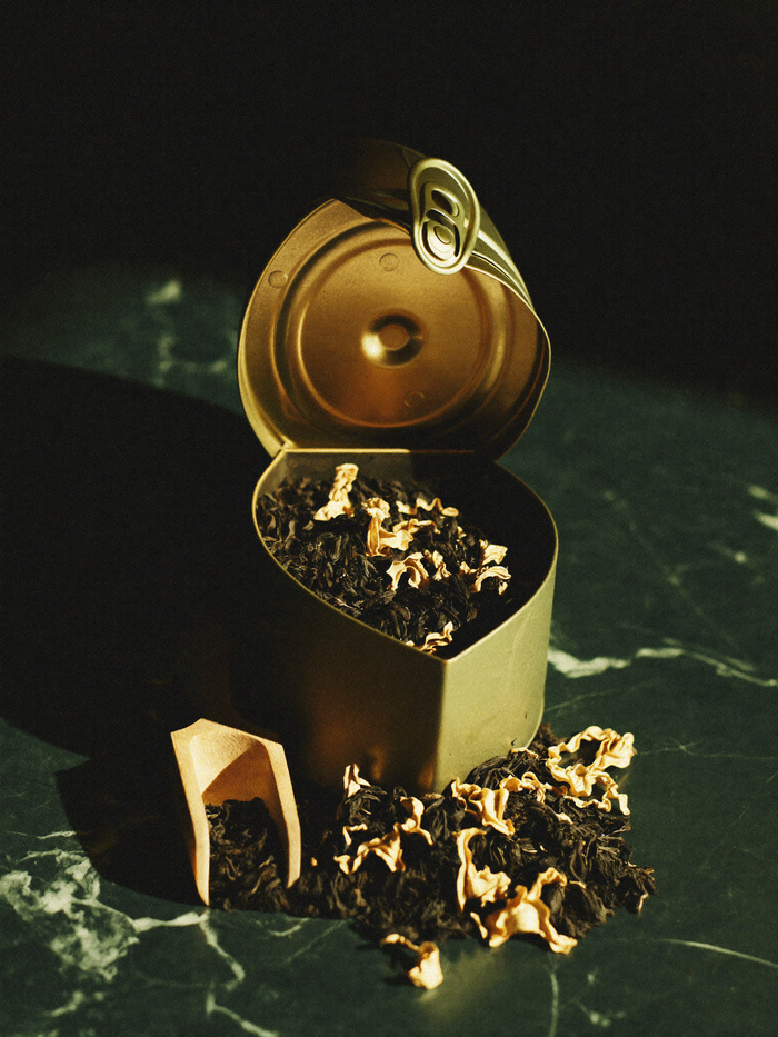 Loose-leaf tea and its package on polished dark marble, lit by warm golden light against a deep emerald-green background falling into shadow.
