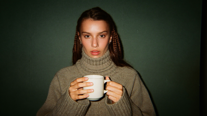 Young woman in a heavy turtleneck sweater holding a plain ceramic mug with both hands against a dark green wall