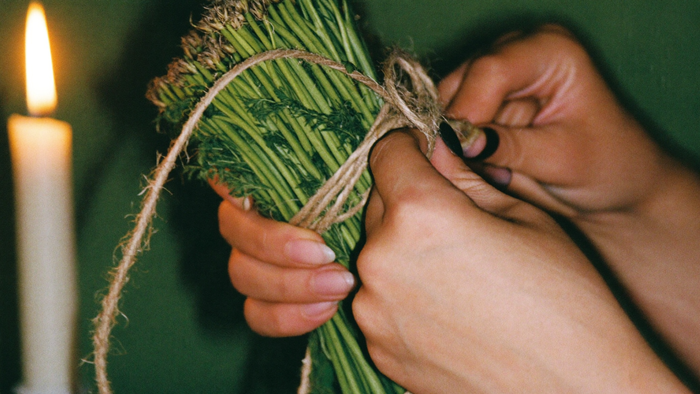 Two hands tying a bundle of freshly cut green herbs with garden twine by candlelight