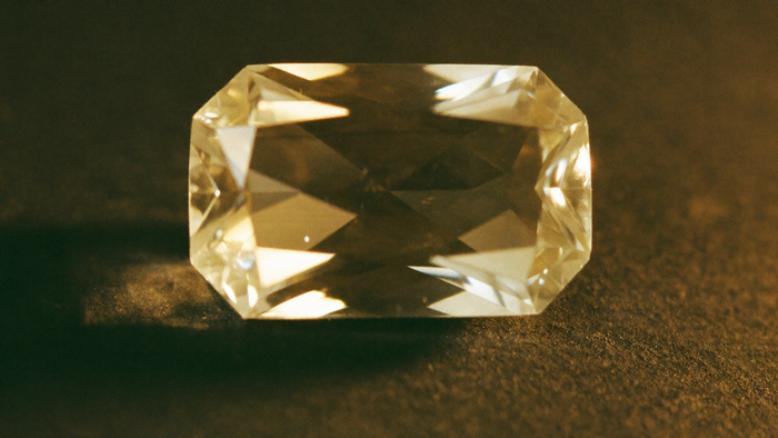 Faceted pale golden topaz on dark slate, lit warmly from one side so the facet edges stay crisp