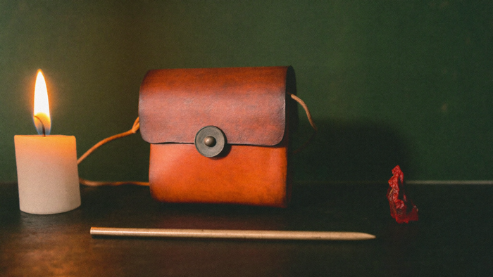 Leather amulet pouch on a cord beside a bronze stylus, red sealing wax and a lit candle on a dark table