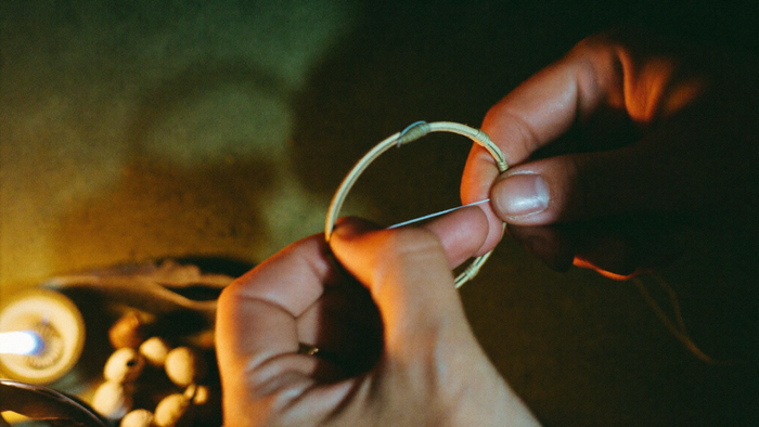 Two hands binding fine thread across a small willow hoop by candlelight, wooden beads and a feather beside them