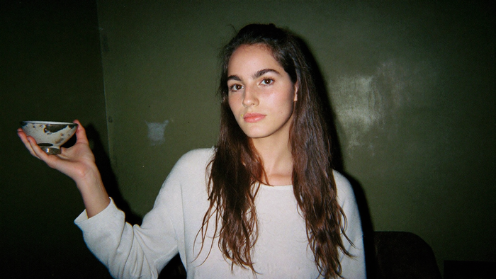 A young woman in a plain sweater holding a small stone offering bowl against a dark green wall