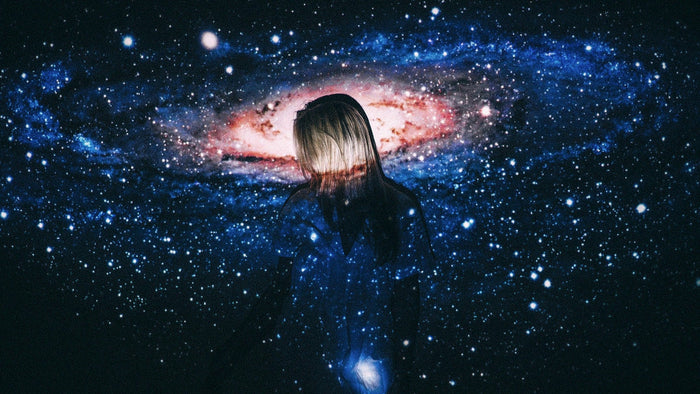 Silhouette of a woman with her head bowed, a spiral galaxy and a field of stars projected across her body in the dark