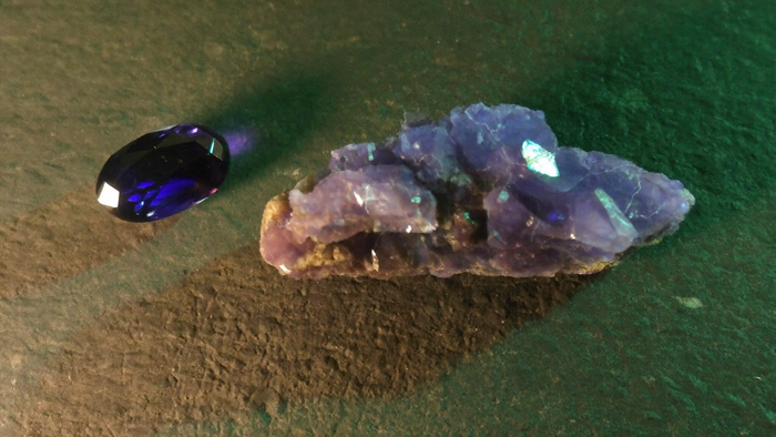 Faceted violet-blue iolite gemstone beside a rough violet cordierite crystal on dark slate in candlelight