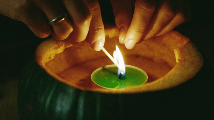 Two hands lighting a small tea light inside a carved pumpkin by candlelight