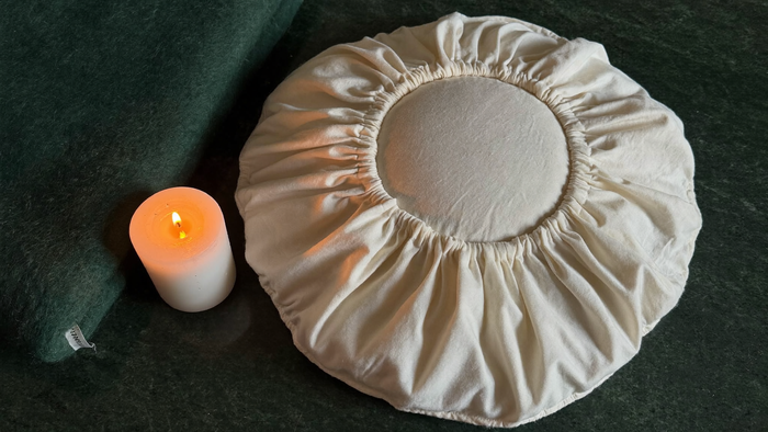Round cotton zafu meditation cushion beside a lit pillar candle and a green wool blanket on a dark surface
