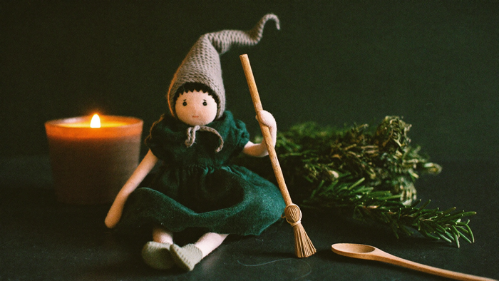 Handmade cloth kitchen witch doll with a straw broom beside dried rosemary, a wooden spoon and a burning candle