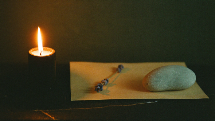 A black pillar candle burning beside a folded paper intention, dried lavender and a smooth pale stone on dark slate