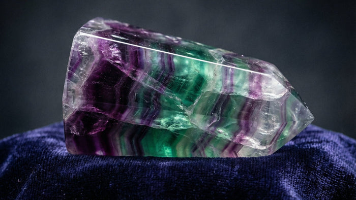 A polished rainbow fluorite point with purple, green and clear bands lying on dark blue velvet against a gray background