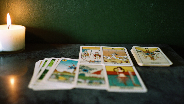 A worn tarot deck separated into small stacks on a dark stone table beside a burning candle