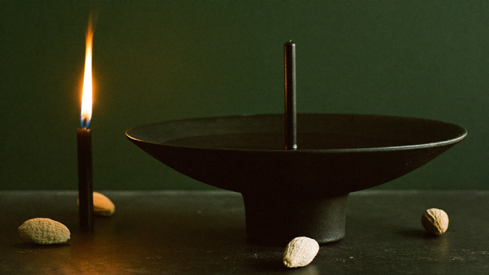 A black bowl of still dark water and an unlit black candle on dark stone, lit by one candle flame