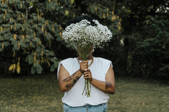 A person in a white tee holds a bouquet of white baby's breath in front of their face, outdoors in soft green light