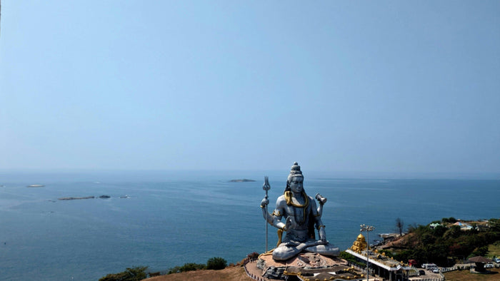 Giant statue of Shiva seated in meditation on a hill above the sea, holding a trident, under a pale blue sky