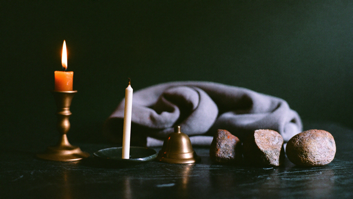 A folded cloth, a cold candle stub in a dish, a brass bell and three stones set aside on a dark altar surface