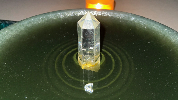 A clear quartz point standing upright in a shallow green bowl of still water, one ripple ring around its base