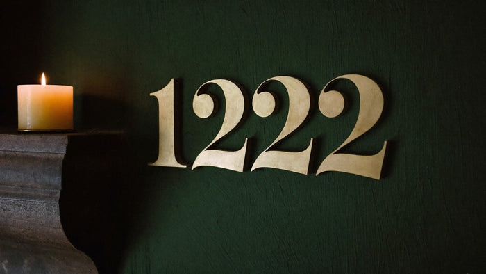 Brass numerals 1222 on a dark green wall beside a lit beeswax candle on a stone ledge, for the 1222 angel number