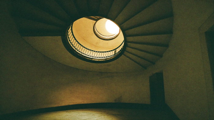 Looking straight up the well of an old spiral staircase, stone flights winding toward a bright round opening high above