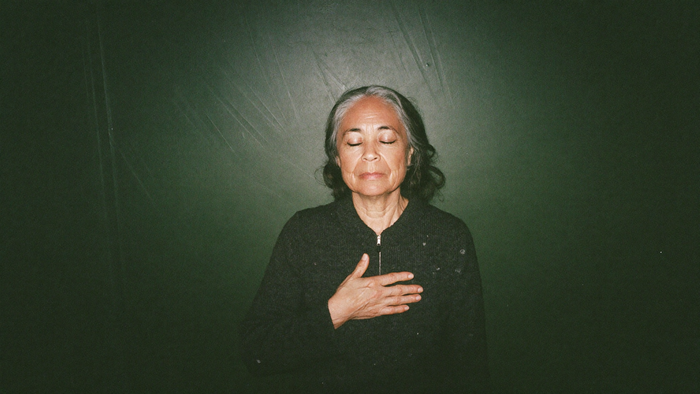 A woman with her eyes closed and one hand resting flat over her heart against a dark green wall