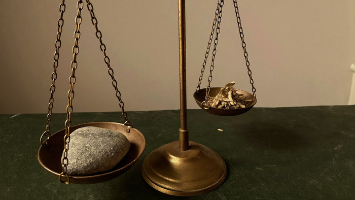 An antique brass balance scale on dark slate, one pan holding a stone and tipping below the other