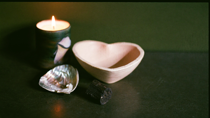 Abalone shell, ceramic smudge bowl and a charcoal disc beside a burning candle on dark stone