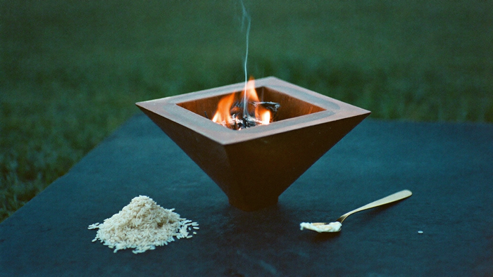 Copper pyramid vessel with a small fire on dark stone at dawn, white rice and a brass spoon of ghee beside it