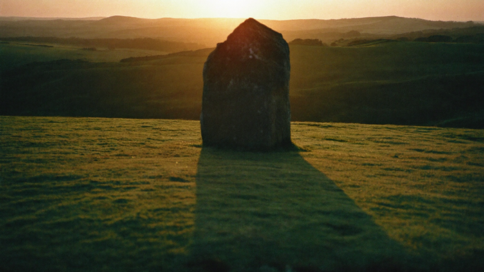 Weathered standing stone on a ridge at dawn, one long straight shadow running across the turf