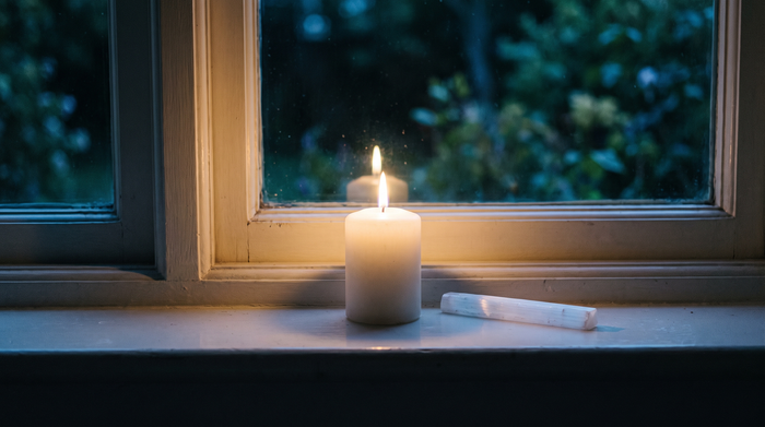 A lit white candle and a pale selenite wand on a windowsill in cool blue night light