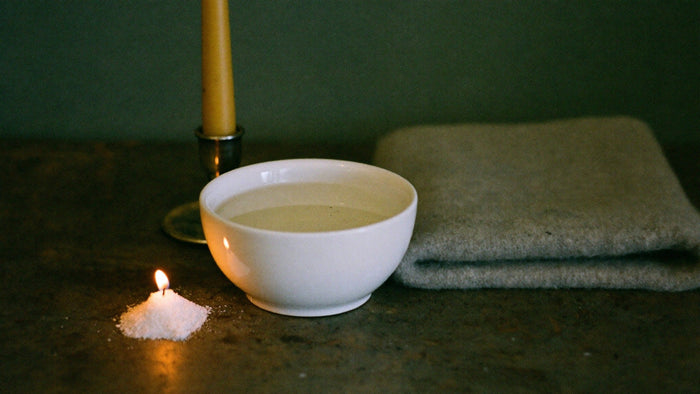 White bowl of water beside a beeswax candle, a small mound of salt and a folded wool blanket on dark stone