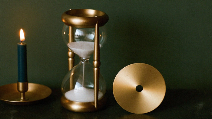Brass hourglass and a plain brass disc beside a lit taper candle against a dark green wall