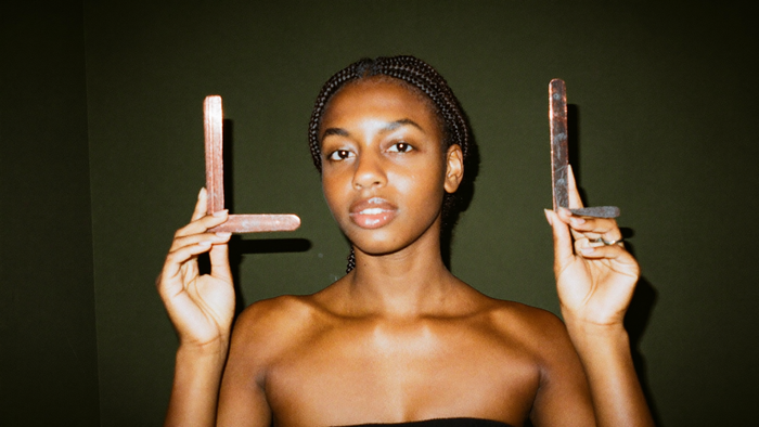 A young woman holding a pair of bent metal dowsing rods upright against a dark green wall