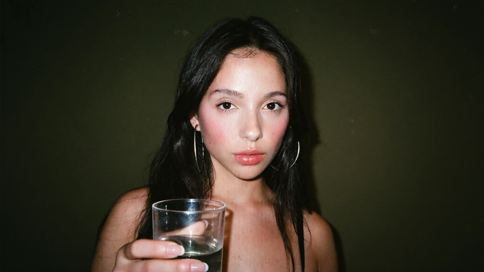 Young woman with dark hair holding a plain glass of water, direct flash, olive green wall behind