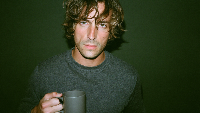 Young man holding a plain grey mug against a dark green wall, lit by direct flash