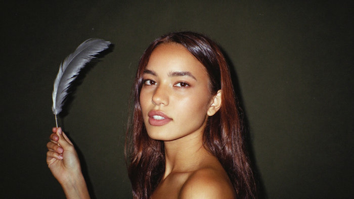 A woman holding a single grey feather upright beside her face against a dark green wall