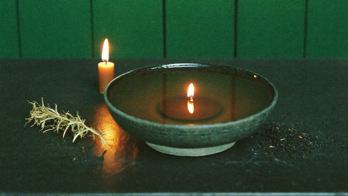 Still water in a dark stone bowl reflecting a single candle flame, dried herbs and dark earth beside it
